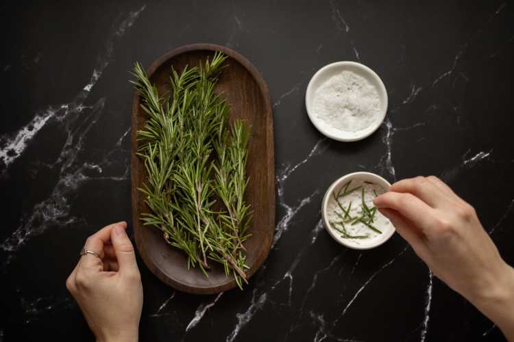 Top view of unrecognizable woman adding fresh twigs of rosemary in bowl with salt for organic scrub placed on black marble table